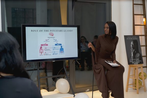 A woman sits beside a presentation screen displaying a diagram in a modern setting.